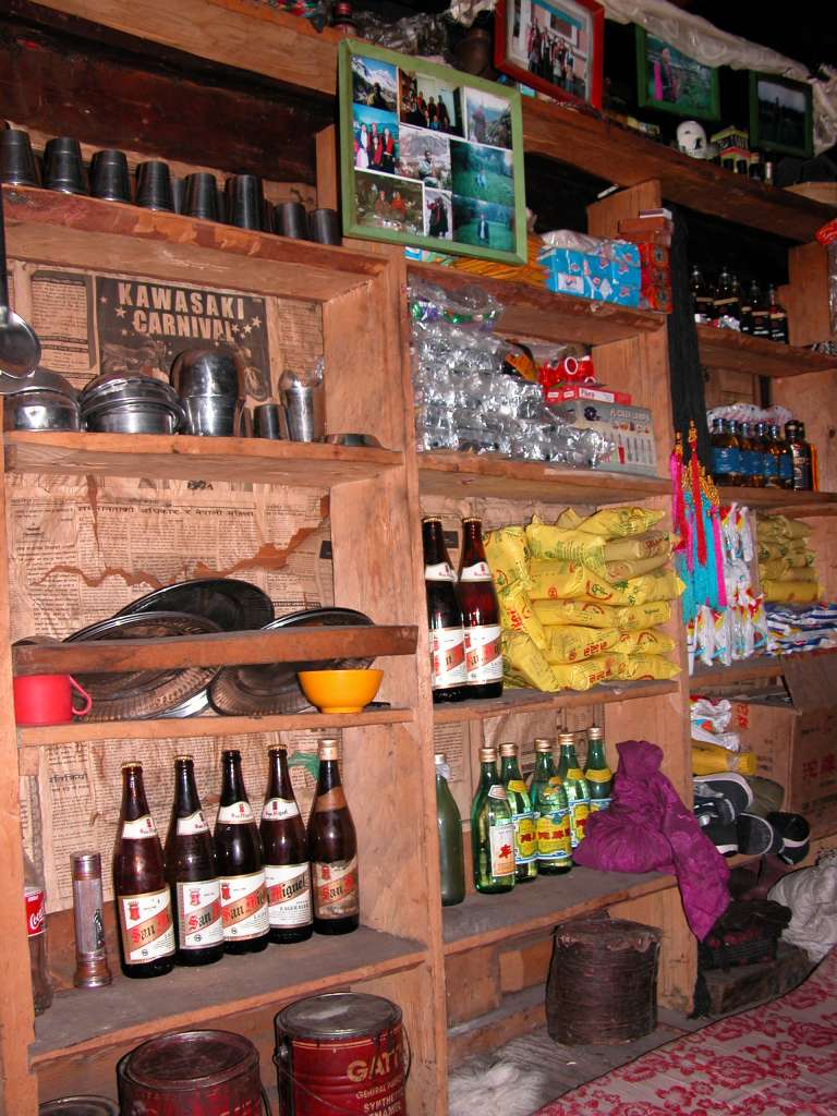 Manaslu 06 11 Syala Lodge Shop The hotel also doubles as a general store. On the left shelves are household cups and bowls,  beer, and paint. In the middle shelves are clothes detergent, cookies, batteries, more beer, butter, Chinese wine, and another can of paint. On the right hand shelves are nail polish, two rows of whisky, noodles, butter, Chinese dish powder, and Goldstar sneakers. Family photos hung proudly near the top.